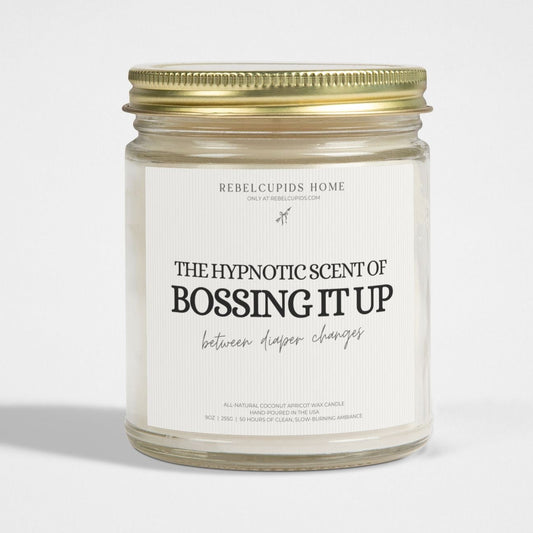 A white candle with the phrase 'THE HYPNOTIC SCENT OF BOSSING IT UP' printed on the jar, along with a gold lid and a wick visible at the top.