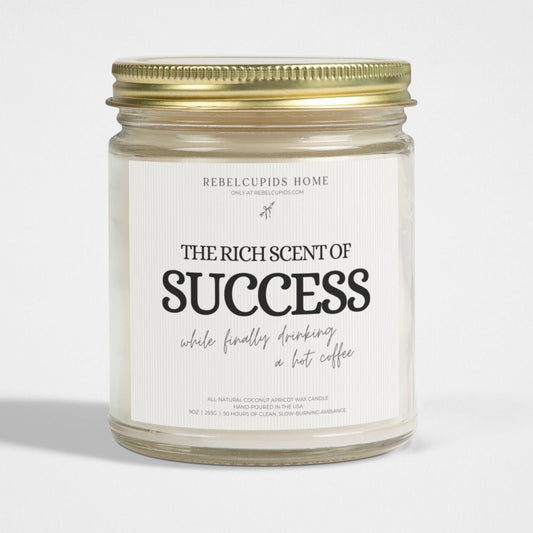 A white candle with a gold lid in a reusable glass vessel, labeled 'The Rich Scents of SUCCESS', with a small amount of wax melted on top.