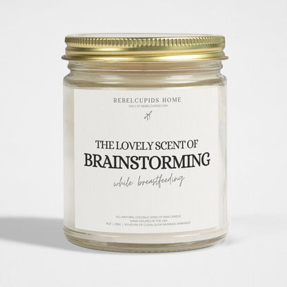 A white candle in a clear glass jar with a gold lid, labeled 'THE LOVELY SCENT OF BRAINSTORMING while breastfeeding'.