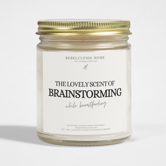 A white candle in a clear glass jar with a gold lid, labeled 'THE LOVELY SCENT OF BRAINSTORMING while breastfeeding'.