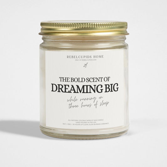 A white candle with the text 'THE BOLD SCENT OF DREAMING BIG while running on three hours of sleep' on its glass vessel, with a gold lid on the side.
