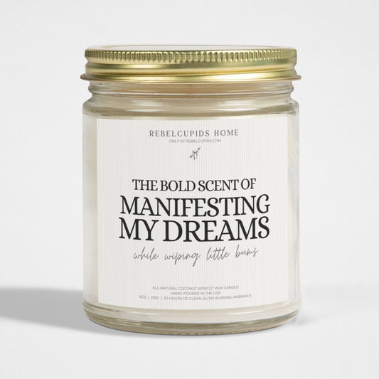 A white candle with the phrase 'THE BOLD SCENT OF MANIFESTING MY DREAMS' printed on it, with a gold lid on the side.