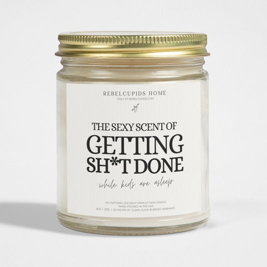 A white candle with the text 'THE SEXY SCENT OF GETTING SH*T DONE' on the label, set in a clear glass jar with a gold lid.
