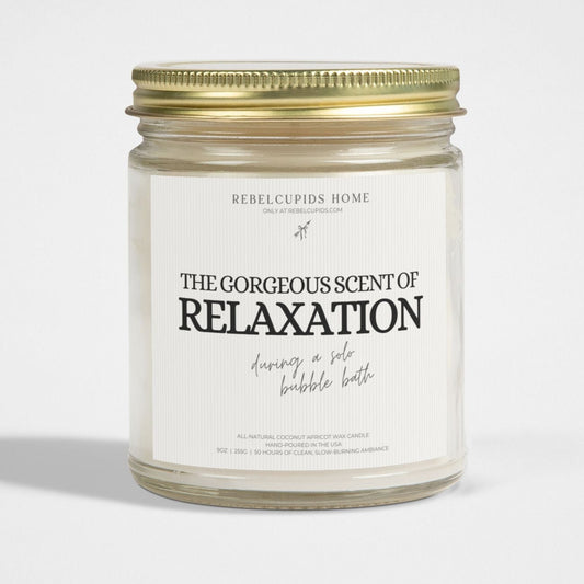 A white candle with the phrase 'THE GORGEOUS SCENT OF RELAXATION' on its label, set in a clear glass jar with a gold lid.