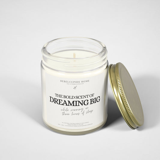 A white candle with the text 'THE BOLD SCENT OF DREAMING BIG while running on three hours of sleep' on its glass vessel, with a gold lid on the side.