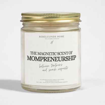 A white candle with a label that reads 'The Magnetic Scent of Mompreneurship', placed in a clear glass jar with a gold lid.