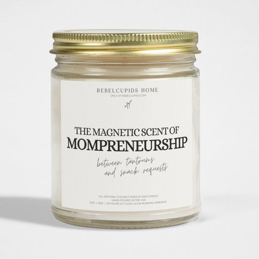 A white candle with a label that reads 'The Magnetic Scent of Mompreneurship', placed in a clear glass jar with a gold lid.